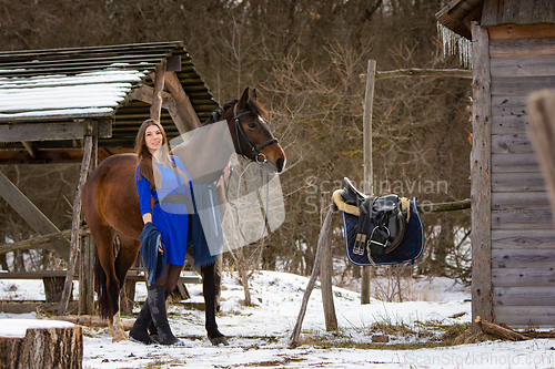 Image of A beautiful girl in a short blue dress walks with a horse near the farm in winter