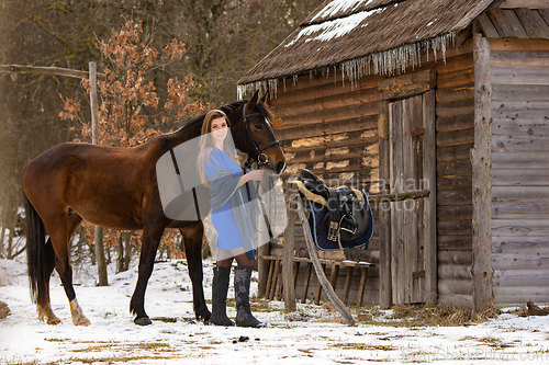 Image of A beautiful girl in a short blue dress stands with a horse from an old wooden house