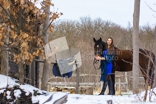 Image of A girl in a blue dress walks with a horse through a farm in winter