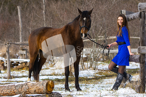 Image of A beautiful girl in a short blue dress walks with a horse at an old farm in winter, the girl smiles and looks into the frame