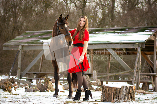 Image of A beautiful girl in a red dress walks with a horse in the rays of the setting sun against the backdrop of old ruins