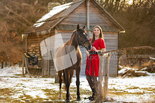 Image of A beautiful girl in a red dress walks through an old farm in the winter in the rays of the setting sun