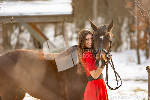Image of A beautiful girl in a red dress hugs a horse in the rays of the setting sun