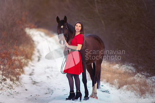 Image of A beautiful girl in a red dress walks with a horse against the backdrop of a winter forest