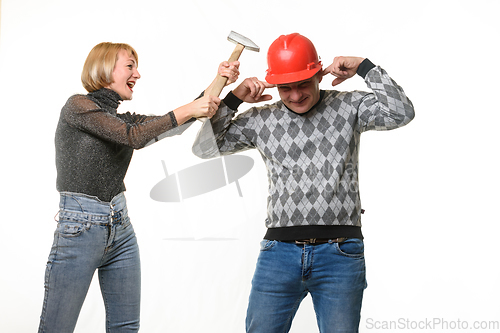 Image of The wife screams and hits her husband on the head with a hammer, the husband is wearing a helmet and he covered his ears with his fingers