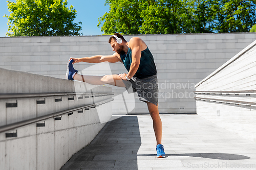 Image of young man in headphones stretching leg outdoors