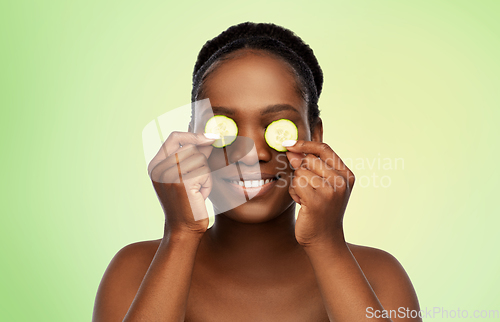 Image of happy african woman making eye mask of cucumbers