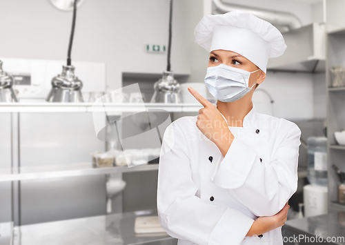 Image of female chef in mask pointing finger up at kitchen