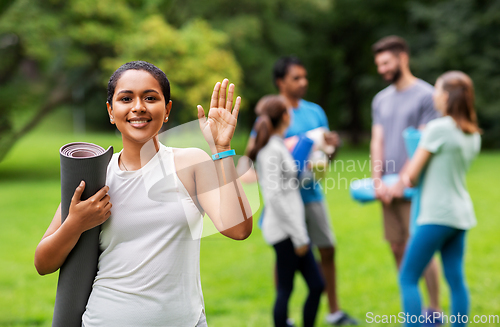 Image of smiling woman with yoga mat waving hand at park