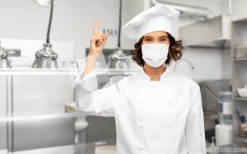 Image of female chef in mask pointing finger up at kitchen
