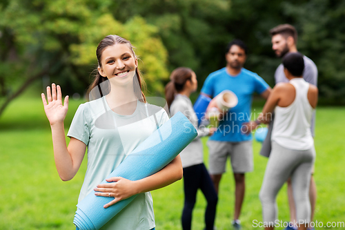 Image of smiling woman with yoga mat waving hand at park