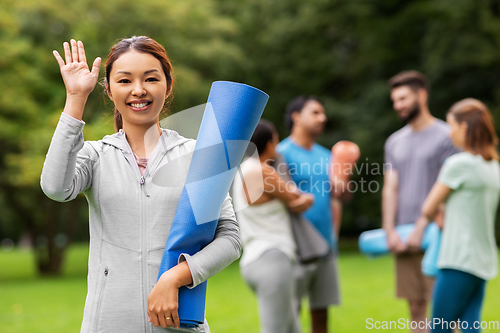 Image of smiling woman with yoga mat waving hand at park