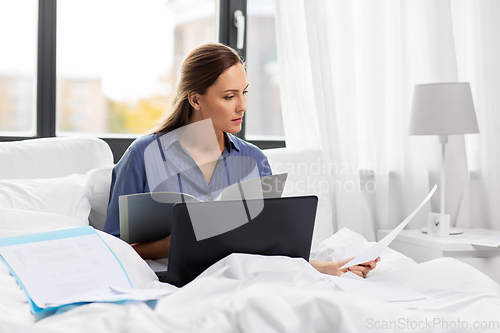 Image of young woman with laptop and papers in bed at home