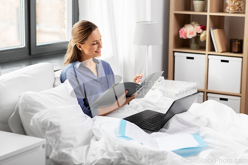 Image of young woman with laptop and papers in bed at home