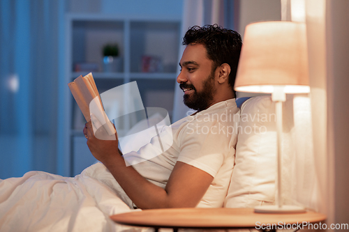 Image of happy indian man reading book in bed at night