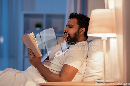 Image of yawning indian man reading book in bed at night