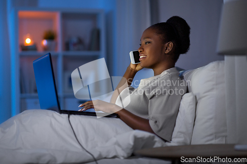 Image of woman with laptop calling on smartphone in bed