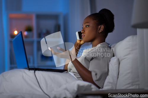 Image of woman with laptop calling on smartphone in bed