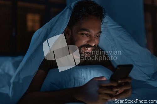 Image of indian man with smartphone in bed at home at night