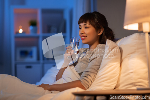 Image of smiling asian woman drinking water at night in bed