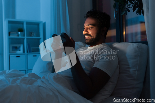 Image of indian man with smartphone in bed at home at night