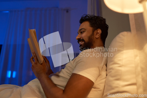 Image of happy indian man reading book in bed at night