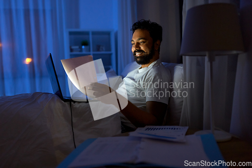 Image of indian man with laptop and papers in bed at night