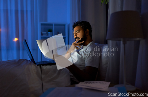 Image of indian man with laptop and papers in bed at night