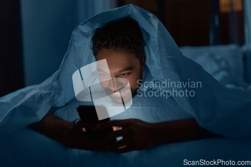 Image of indian man with smartphone in bed at home at night