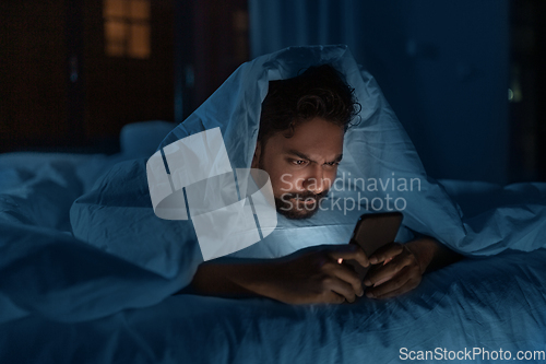 Image of indian man with smartphone in bed at home at night