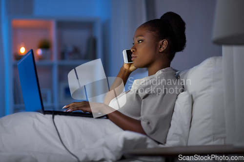 Image of woman with laptop calling on smartphone in bed