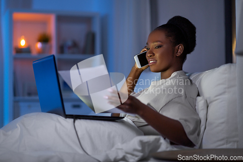 Image of woman with laptop calling on smartphone in bed