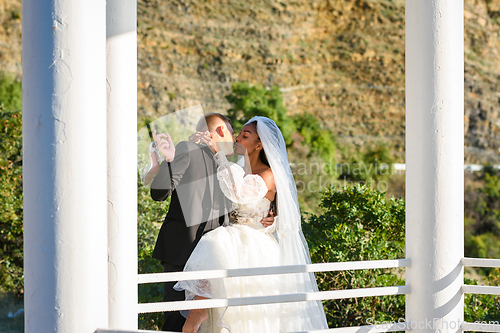 Image of Young beautiful interracial newlyweds are kissing in the gazebo, against the backdrop of sunny mountains