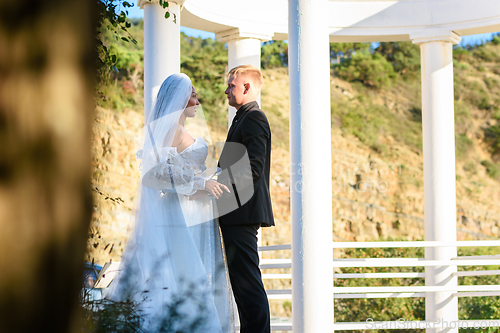 Image of Young beautiful interracial newlyweds lovingly look at each other against the background of white columns