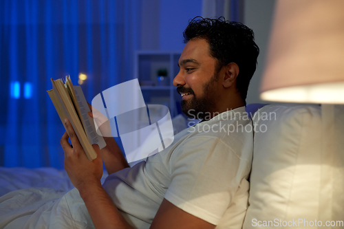Image of happy indian man reading book in bed at night