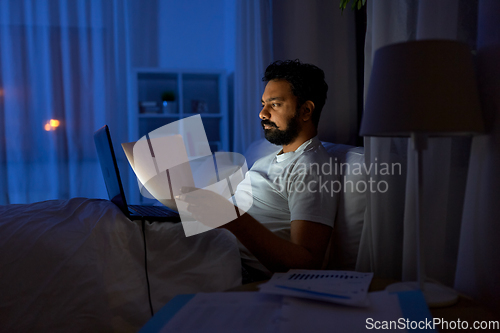 Image of indian man with laptop and papers in bed at night