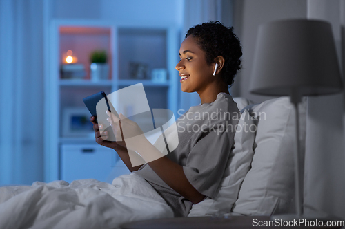 Image of woman with tablet pc in earphones in bed at night