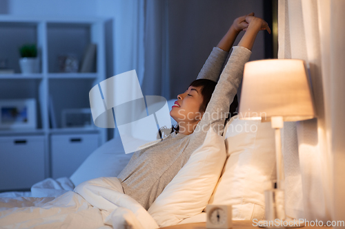 Image of tired asian woman stretching in bed at night