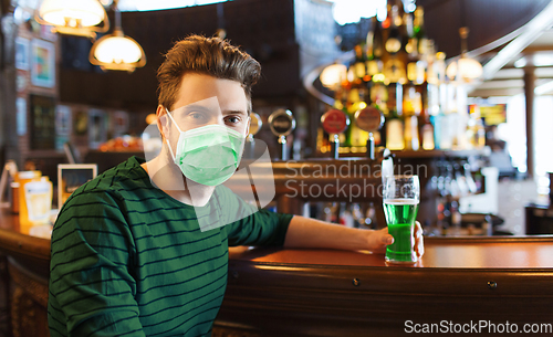 Image of young man in mask drinking green beer at bar