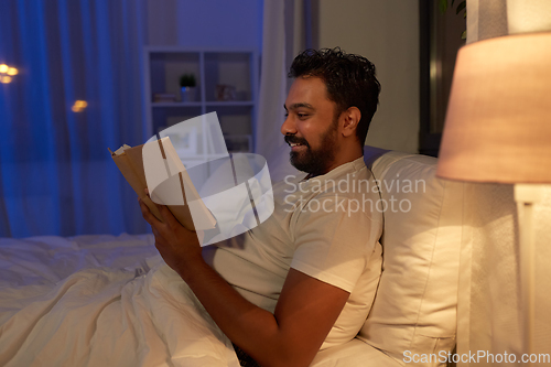 Image of happy indian man reading book in bed at night