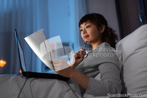 Image of asian woman with laptop and papers in bed at night