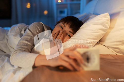 Image of sleepy asian woman with clock in bed at night