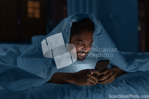 Image of indian man with smartphone in bed at home at night