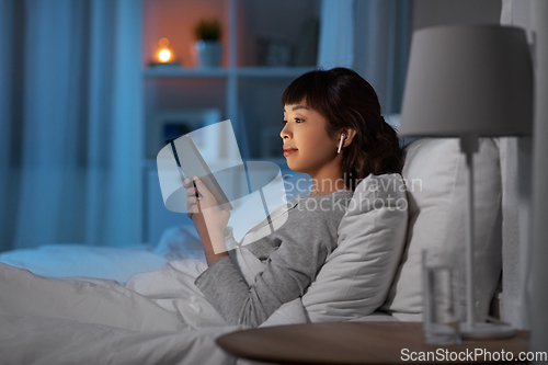 Image of woman with tablet pc in earphones in bed at night