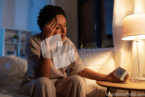 Image of african woman with clock sitting on bed at night