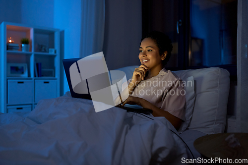 Image of smiling woman with laptop in bed at home at night