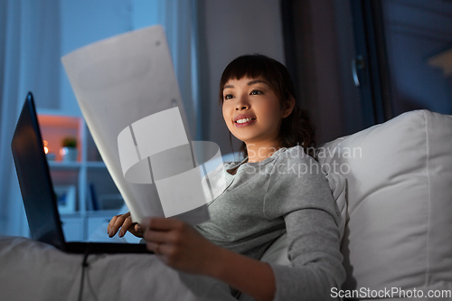 Image of asian woman with laptop and papers in bed at night