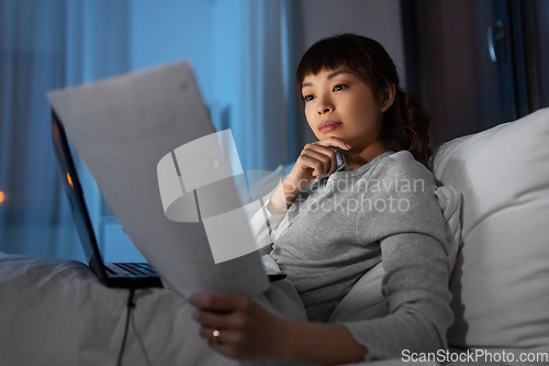 Image of asian woman with laptop and papers in bed at night