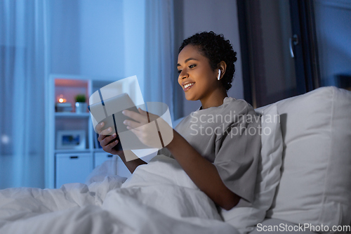 Image of woman with tablet pc in earphones in bed at night