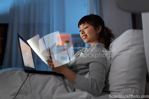 Image of asian woman with laptop and papers in bed at night